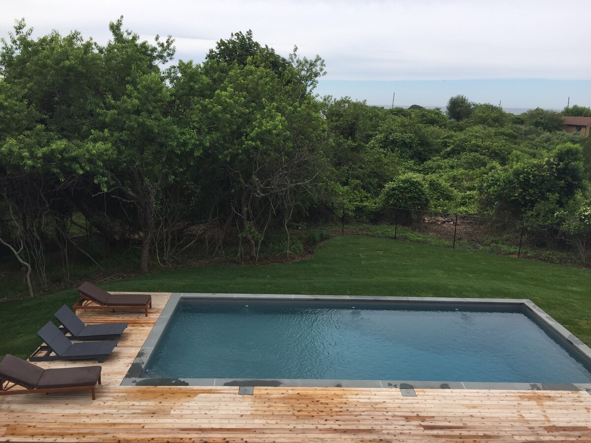A serene view of an in-ground pool, surrounded by wooden decking. Two lounge chairs are positioned for relaxation beside the water. Lush greenery and trees create a natural backdrop, providing privacy and a tranquil atmosphere.