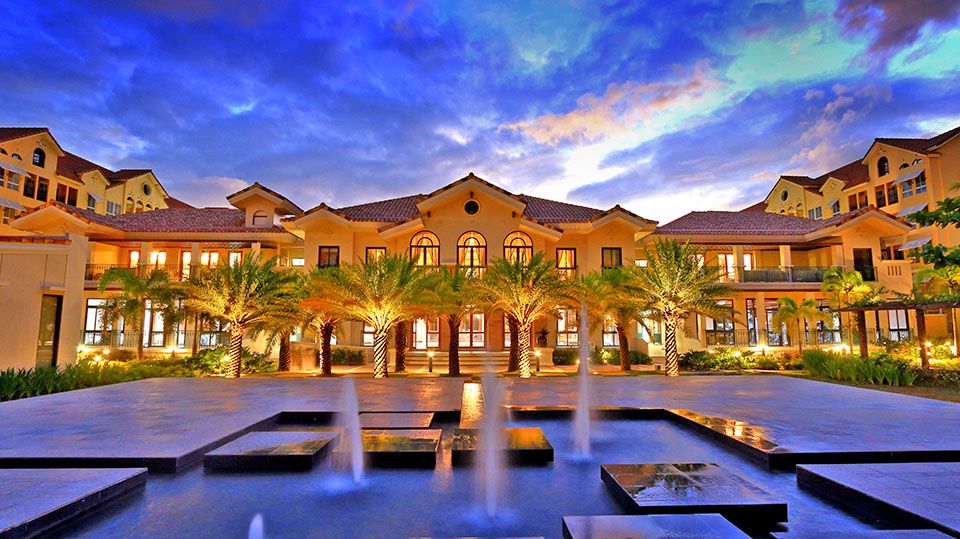 A Mediterranean-inspired building is showcased with palm trees flanking a central fountain. The architecture features arched windows and a tiled roof, with a vibrant sky providing a dramatic backdrop. Multiple levels and balconies are visible, creating an inviting courtyard atmosphere.