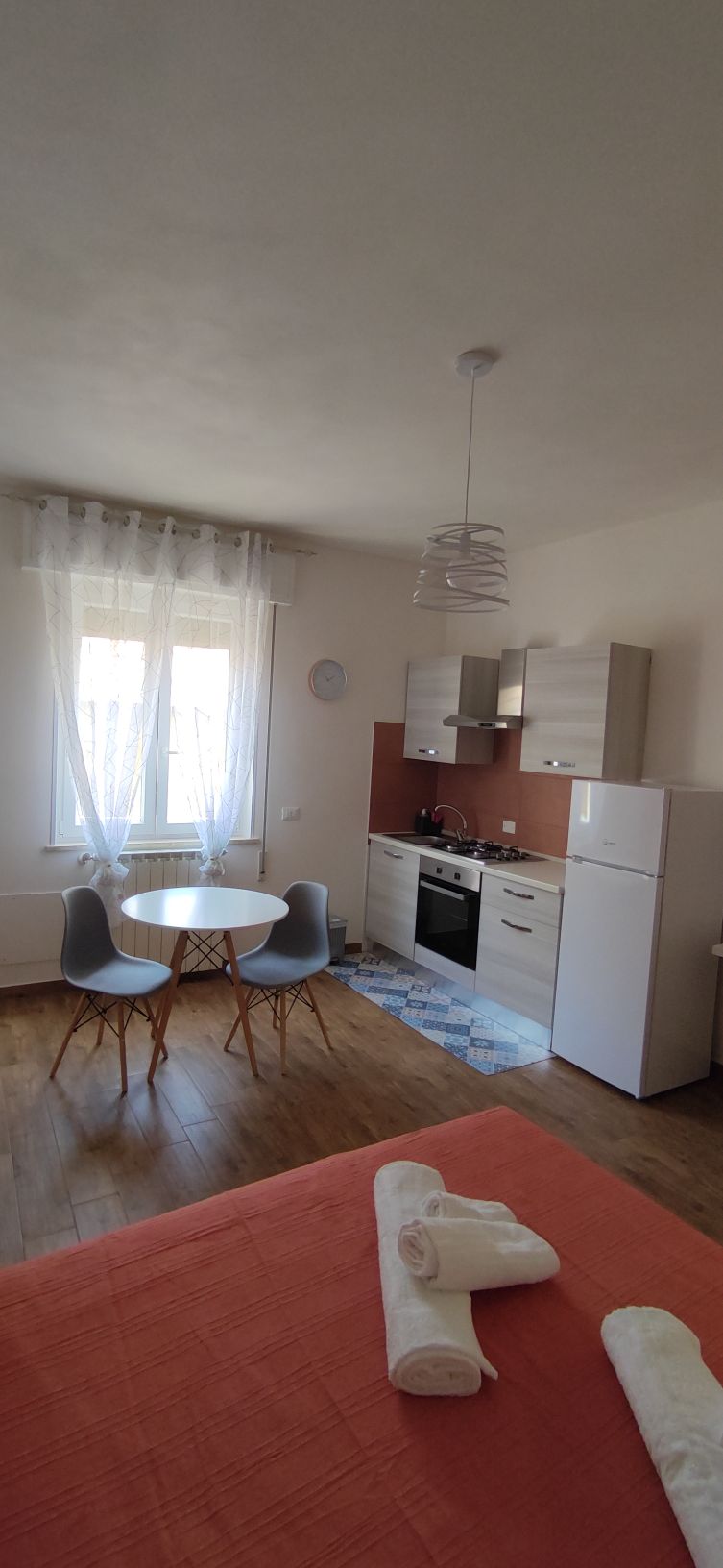 A well-lit studio apartment features a kitchenette with modern appliances, including a refrigerator and stove. A small dining table with two chairs is positioned by the window, allowing natural light to fill the space. The flooring is finished in warm wood tones, enhancing the overall comfort.