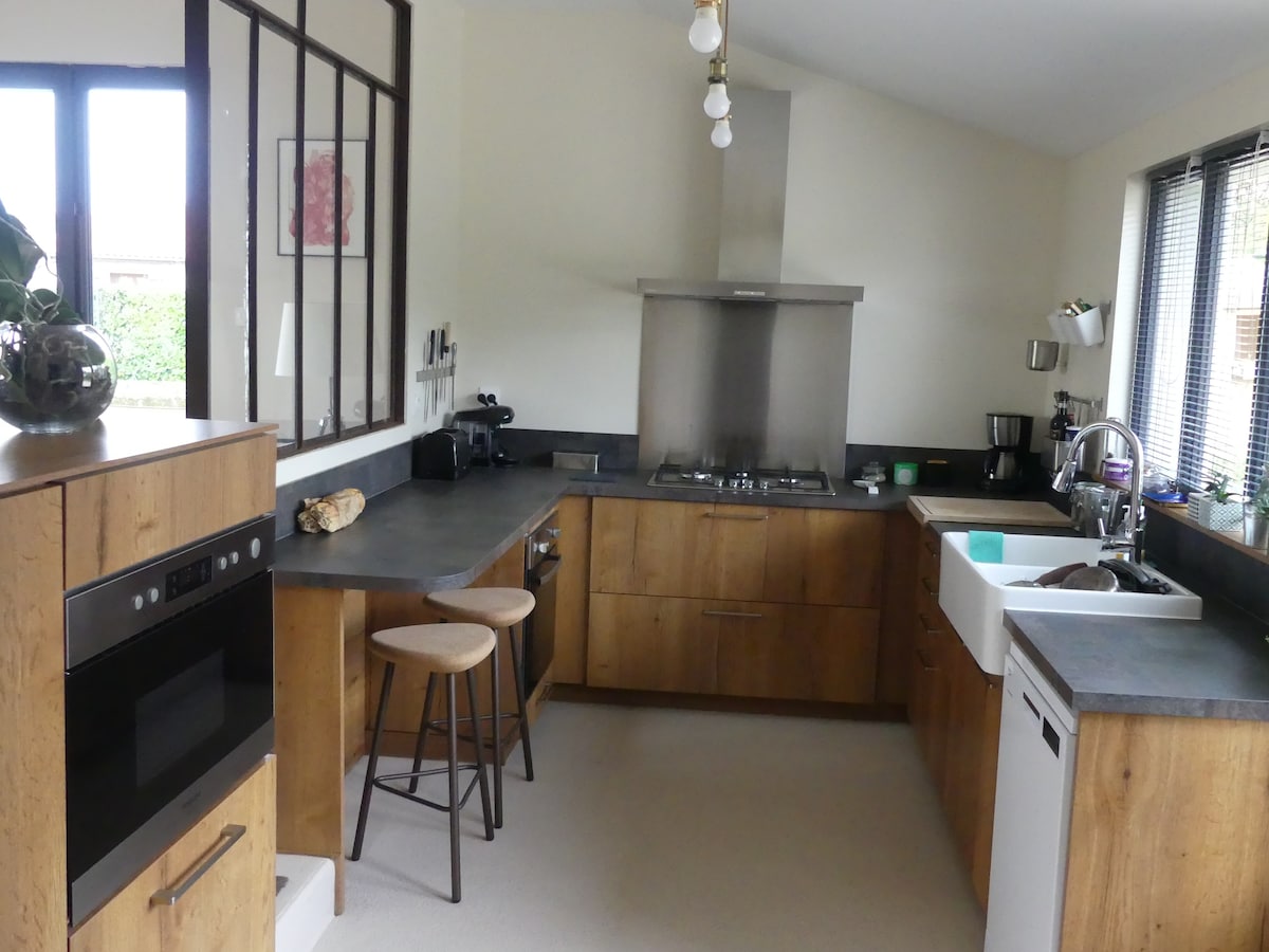 A modern kitchen features wood cabinetry and countertop, complemented by stainless steel appliances. Two bar stools are positioned at a counter, while a gas stove is located against the far wall. Natural light filters through large windows, enhancing the open feel of the space.