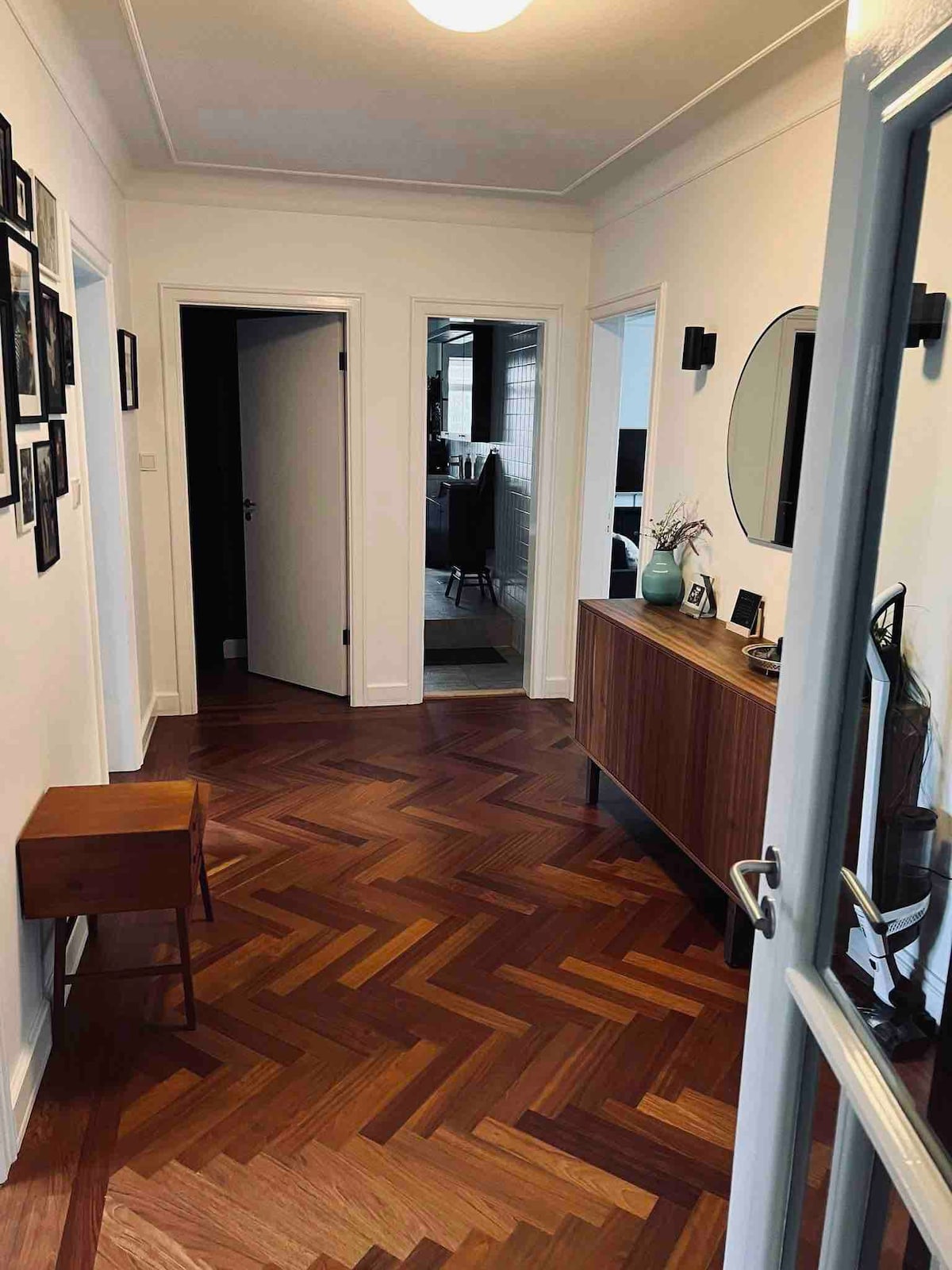 A hallway is shown, featuring herringbone-patterned wooden flooring. Light walls enhance the spaciousness. A wooden console table is positioned against one wall, adorned with decorative items. Framed photographs line the adjacent walls. Doorways lead to other rooms, adding depth to the layout.