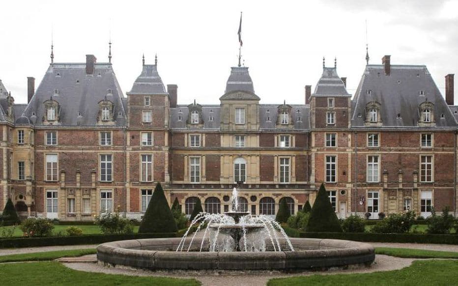 A large chateau features an intricate facade with various architectural elements. A fountain is positioned prominently in the foreground, surrounded by neatly trimmed hedges and landscaped gardens. The building's multiple roofs and windows reflect historical design, emphasizing its grandeur and elegance.