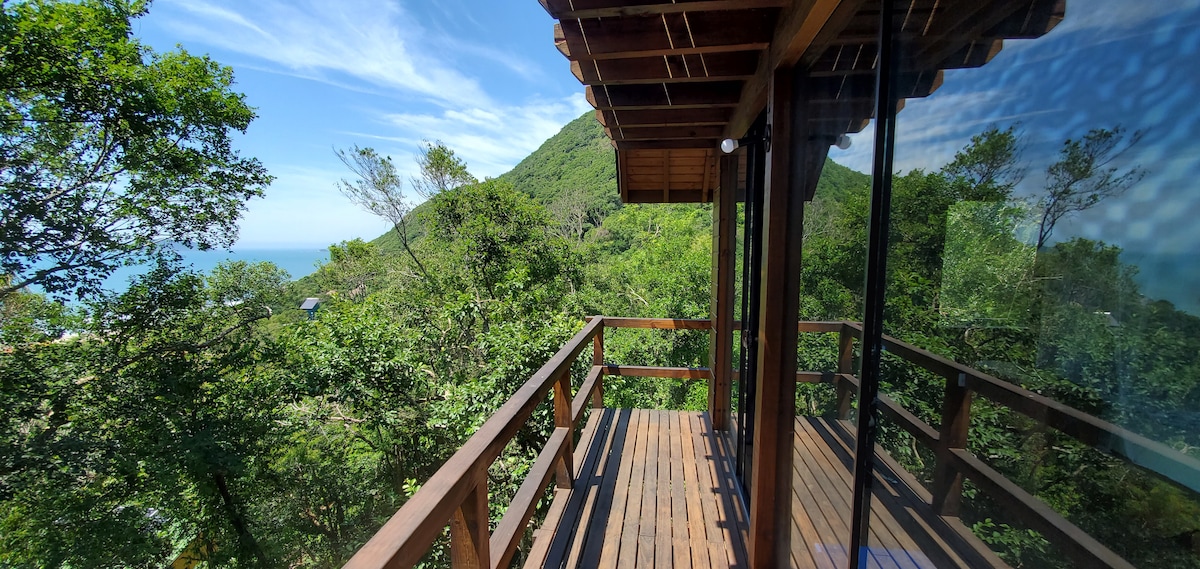 A wooden balcony extends from the house, framed by lush greenery and offering a view of the surrounding hills and ocean. Expansive glass doors allow natural light to fill the space, creating a seamless connection between indoor and outdoor environments.