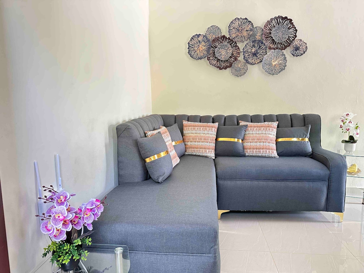 A modern gray sectional sofa is positioned against a light-colored wall, adorned with decorative floral wall art. Soft pillows in varying patterns enhance the seating area. A small glass table displays an arrangement of purple flowers, adding a touch of color to the space.