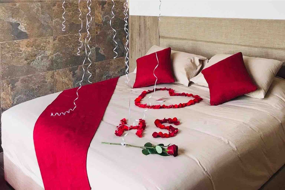 A spacious bed is adorned with a soft beige cover, complemented by two red pillows. Rose petals are arranged in a heart shape on the bed, accompanied by a single rose. Red ribbons float gently above, adding a touch of elegance to the atmosphere.
