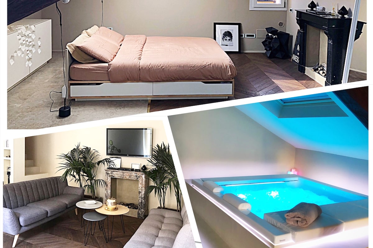 The image displays a serene master suite featuring a spacious king-size bed with a neutral-toned duvet. A cozy sitting area is equipped with soft gray sofas and a marble fireplace. A sleek hot tub with soft blue lighting creates a relaxing atmosphere.