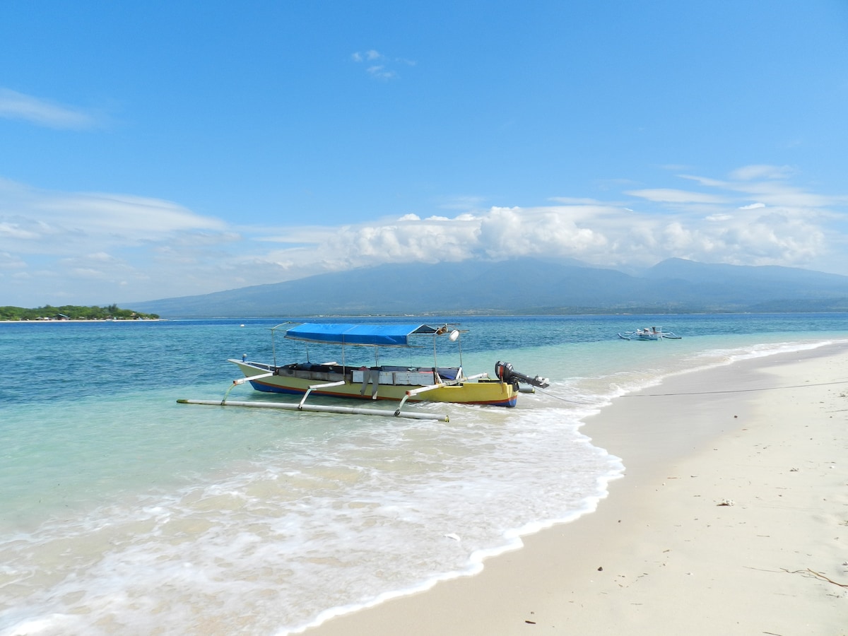 A traditional boat rests on the sandy shore, with shallow turquoise waters gently lapping at its sides. The backdrop features a clear blue sky scattered with clouds and a distant view of a mountainous landscape, enhancing the tranquil island atmosphere.
