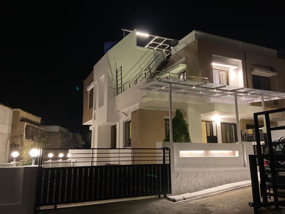 The exterior of the property showcases a modern two-story building illuminated at night. A covered entryway is visibly highlighted, and a staircase is seen along one side. Surrounding greenery and well-placed lighting enhance visibility and accessibility.