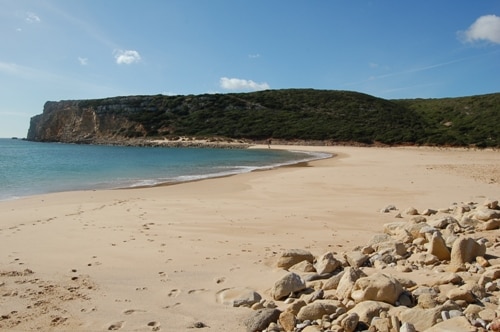 A serene beach scene is captured, featuring a wide sandy shore bordered by gentle waves. The coastline is accentuated by a rocky outcrop and lush green hills in the background. Light clouds float in a blue sky, complementing the tranquil atmosphere.