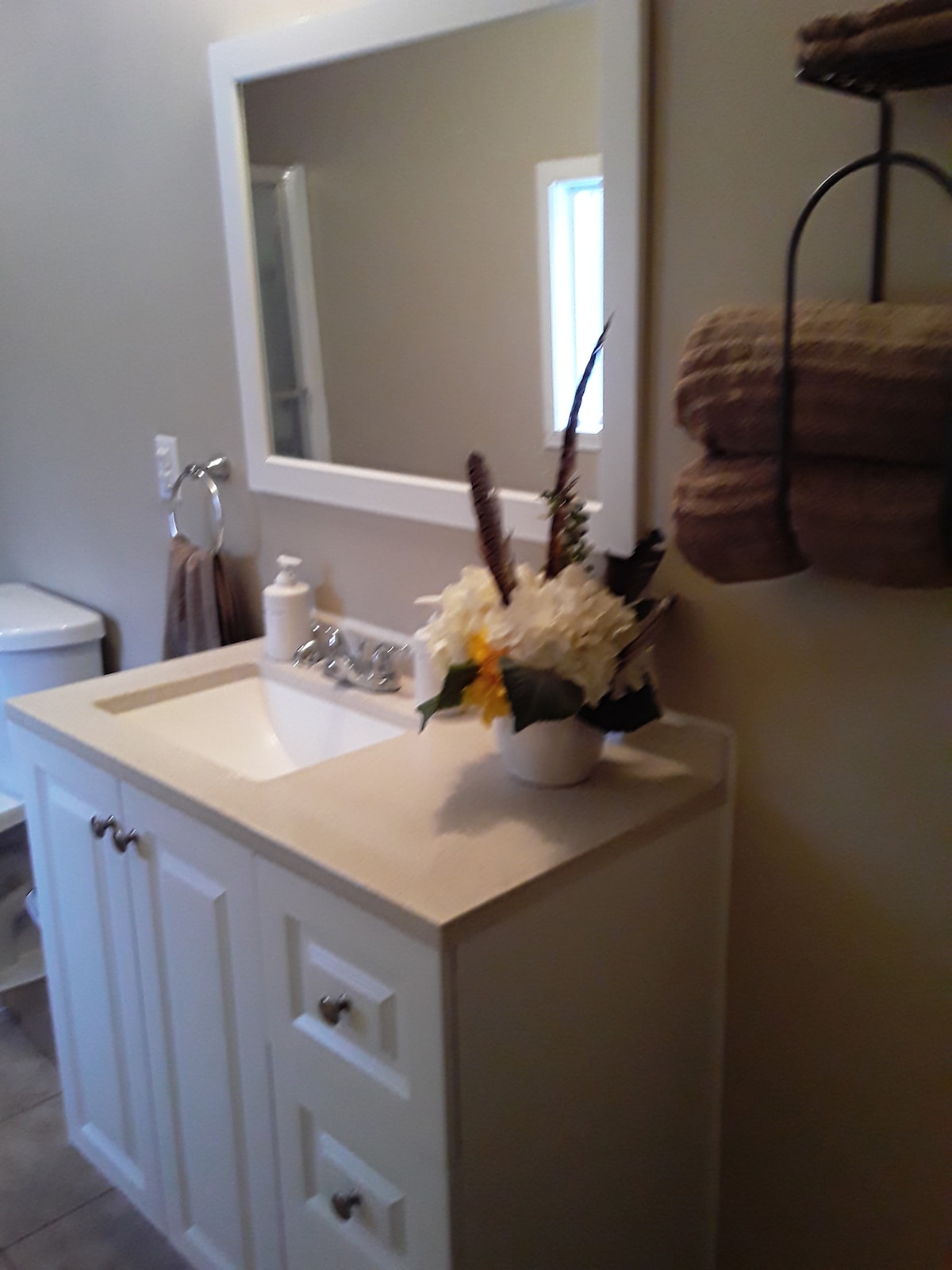 A bathroom is shown with a white vanity, featuring two drawers and a sink. A large mirror hangs above the sink. A decorative arrangement of flowers is placed on the counter, alongside a soap dispenser. Fresh towels are neatly hung from a wall-mounted rack.