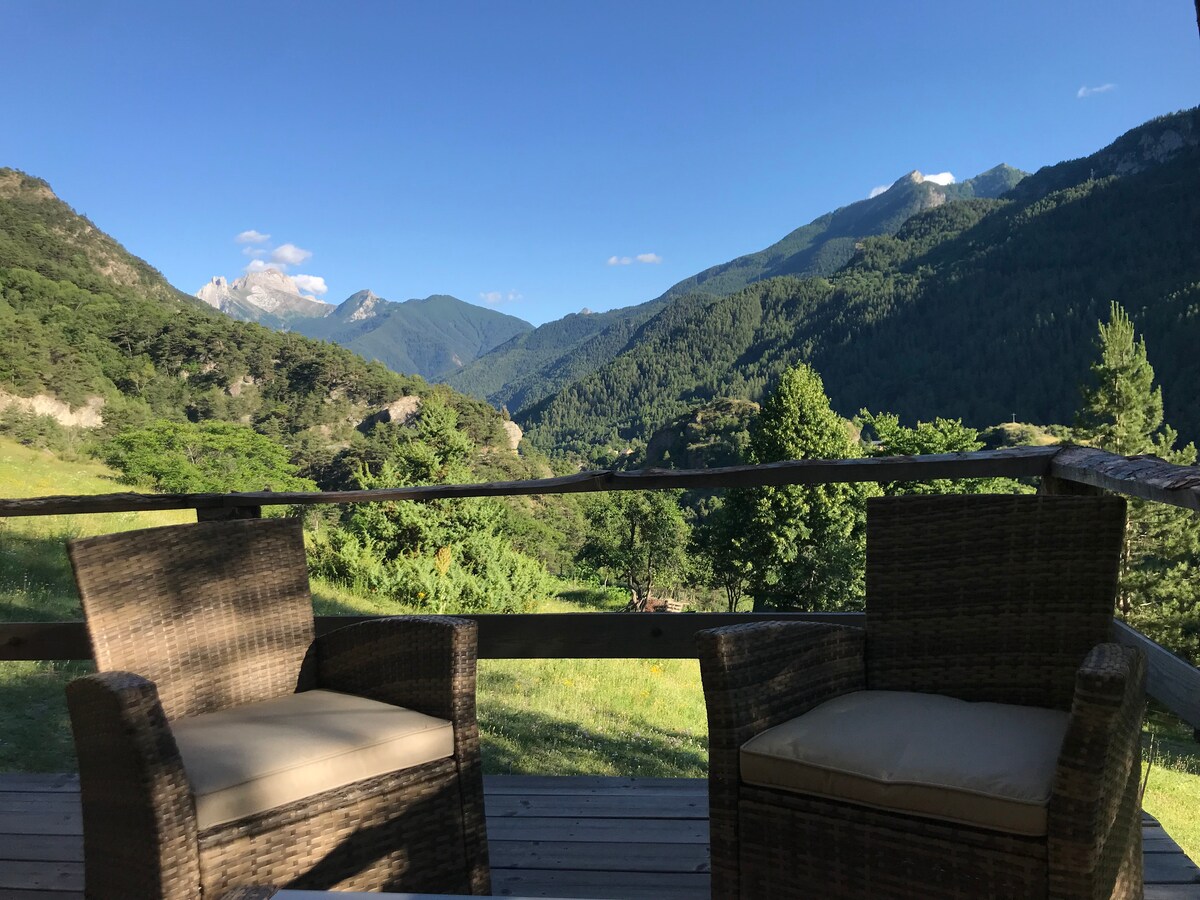 Two wicker chairs with soft cushions are positioned on a wooden terrace, offering an unobstructed view of rolling mountains and lush greenery. The bright blue sky is visible in the background, enhancing the serene natural surroundings.