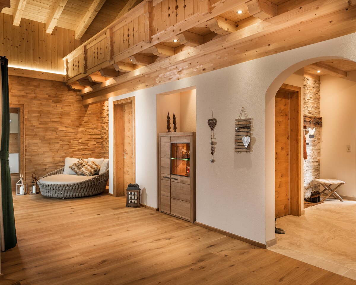 An interior space showcases a combination of wooden beams and stone accents. A round seating area is positioned near a wall, complemented by natural light flowing from a nearby doorway. Decorative items are placed on shelves, enhancing the warm ambiance.