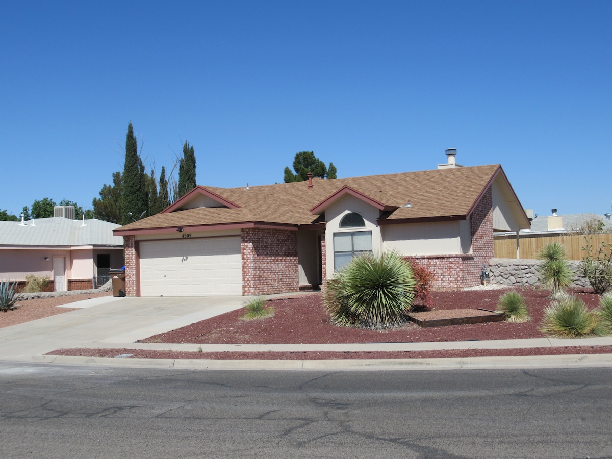 A single-story home is displayed with a brick facade and a gabled roof. Desert landscaping features shrubs and ornamental grasses, complementing the neutral exterior. A paved driveway leads to a two-car garage, and a boundary fence offers privacy.