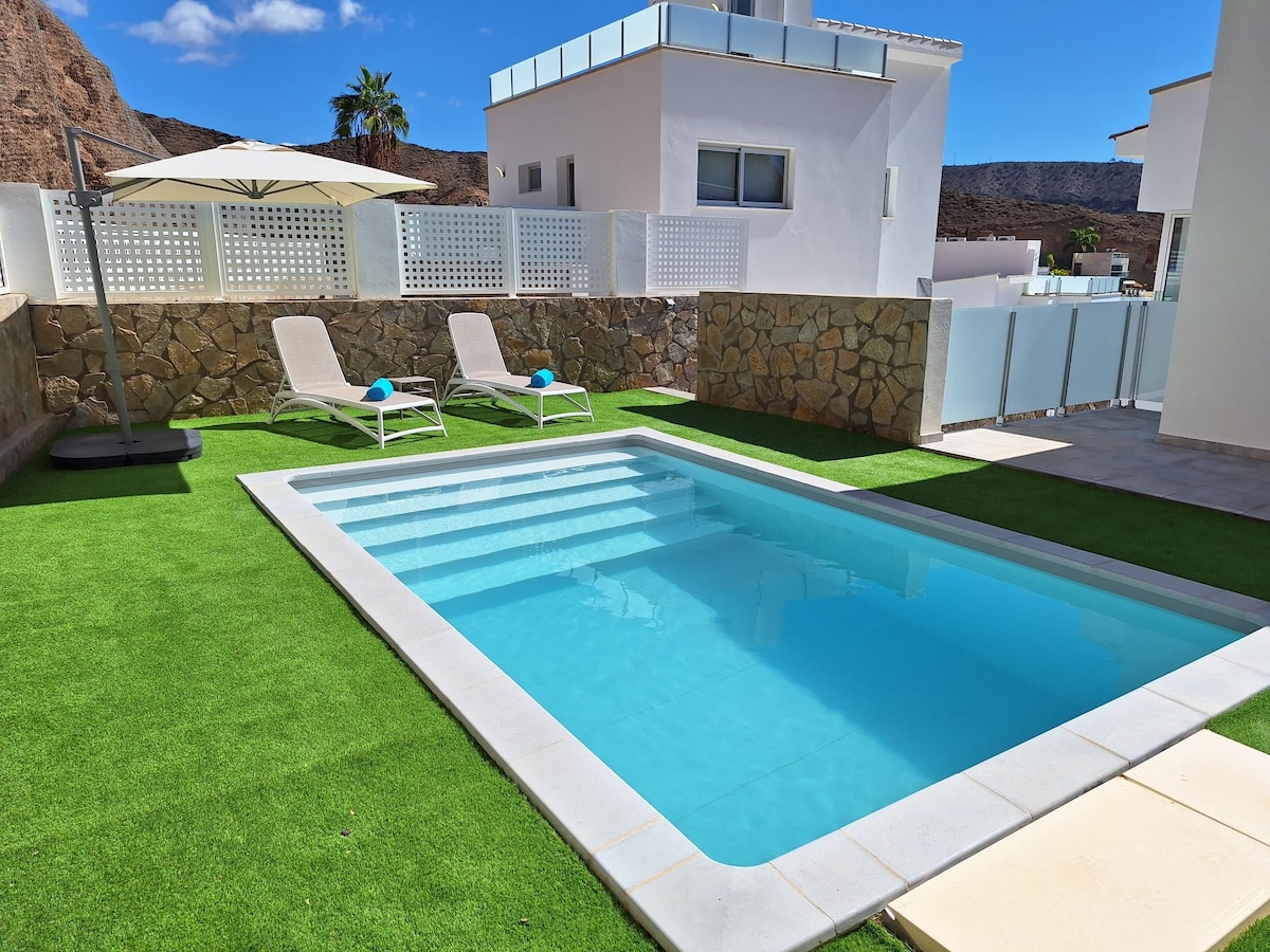 A private pool is featured within a neatly landscaped area, surrounded by a stone wall. Two loungers with blue cushions are positioned nearby, complemented by a large umbrella providing shade. Lush green grass enhances the inviting setting, with a clear blue sky above.