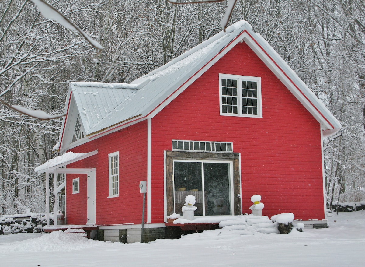 A charming red barn-style house is surrounded by a snowy landscape, highlighting its rustic design. Large windows offer glimpses of the inviting interior, while a covered porch with two white chairs provides a cozy outdoor seating area. The scene reflects the peaceful winter setting.