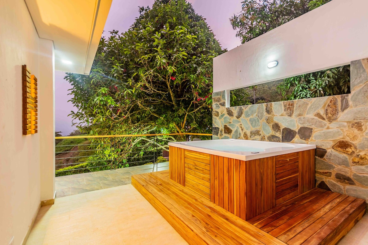An outdoor jacuzzi is set on a wooden deck surrounded by stone walls and lush greenery. Soft lighting highlights the area, creating a serene atmosphere. The view encompasses the natural landscape, enhancing the tranquil setting for relaxation.