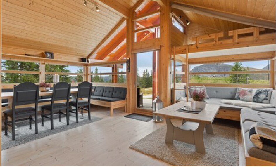 An open-plan living space features high wooden ceilings and large windows that provide views of the surrounding landscape. A modern dining table with six black chairs is positioned beside a cozy seating area. A soft rug covers the wooden floor, adding comfort to the room.