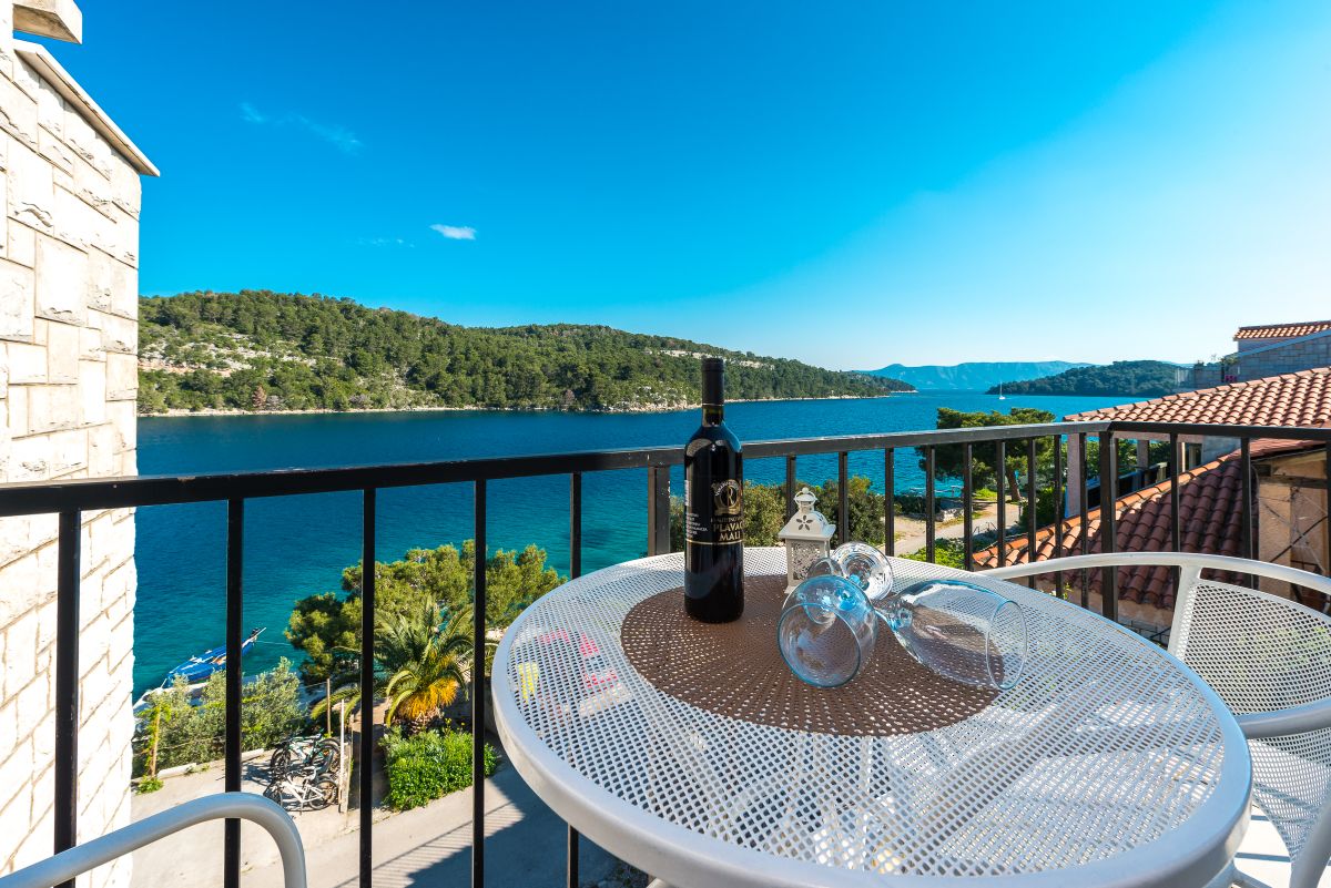A balcony view overlooking a calm bay is showcased, featuring a round table with a bottle of wine and two glasses. Surrounding greenery and distant hills are visible under a clear blue sky, creating a serene coastal atmosphere.