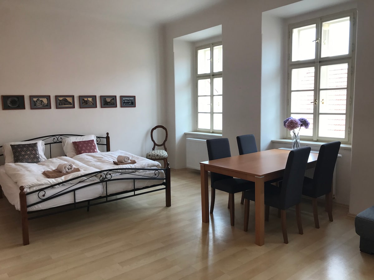 A spacious room features a double bed with light bedding and towels neatly arranged. A wooden dining table is accompanied by four chairs, and two large windows provide natural light. Framed artwork adorns the wall, enhancing the room's serene environment.