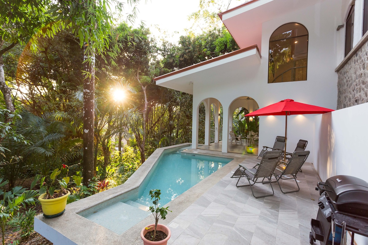 An inviting outdoor area features a swimming pool surrounded by lush greenery. A red umbrella offers shade over a set of lounge chairs, while sunlight filters through the trees, creating a serene atmosphere. A small potted plant adds a refreshing touch.