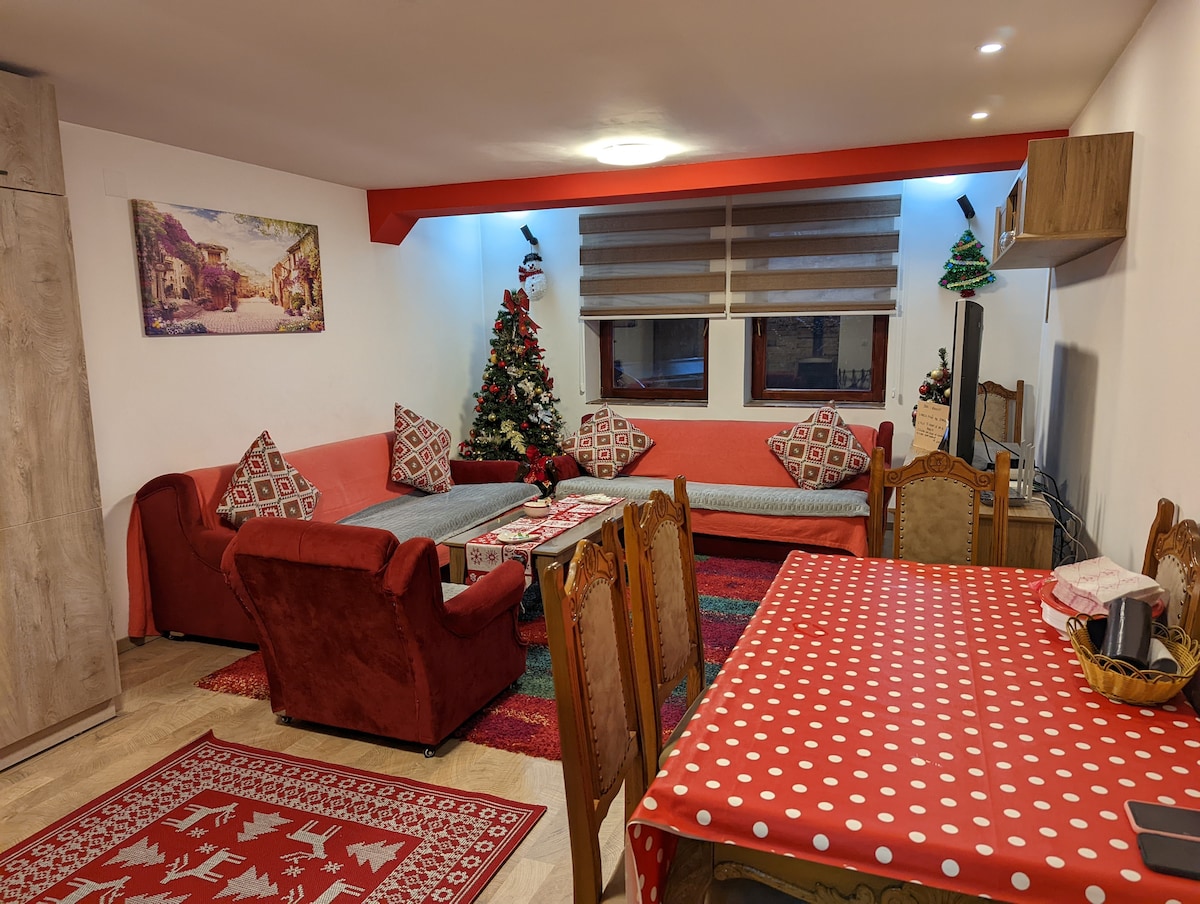 A living area features a cozy arrangement of seating, including a red armchair and a sofa adorned with patterned cushions. A festive Christmas tree is positioned in the corner, while a dining table with a polka dot tablecloth complements the warm, inviting setting.