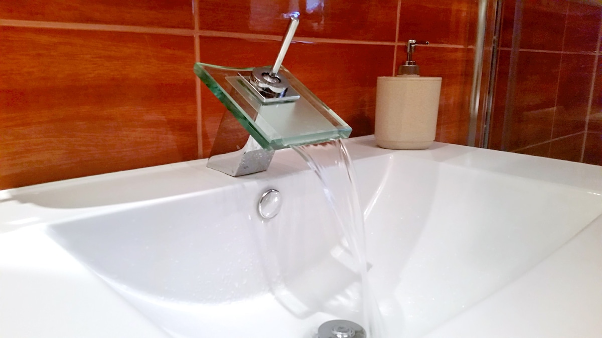A modern bathroom features a sleek, glass and metal faucet pouring water into a white, deep bathtub. The rich brown tiles create a warm backdrop, while a minimalist soap dispenser is positioned nearby, adding a touch of elegance to the space.