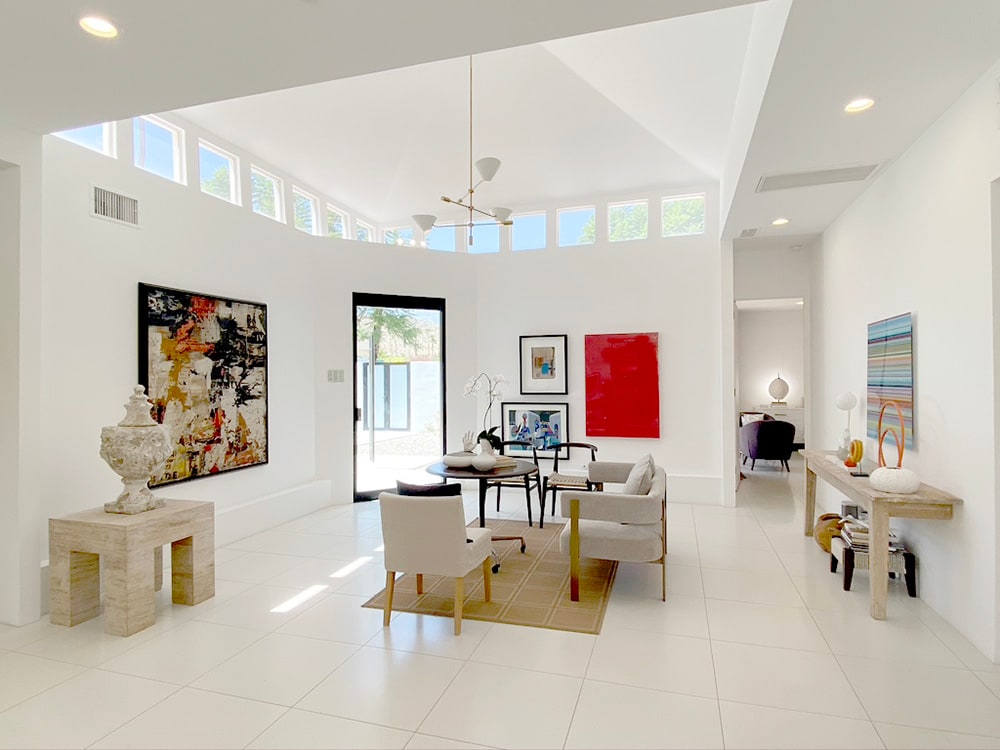 An open living area features a large circular ceiling with multiple windows allowing ample natural light. The space is furnished with a small dining table, chairs, and stylish décor, complemented by vibrant wall art. A glass door provides an entryway to the outdoor area.