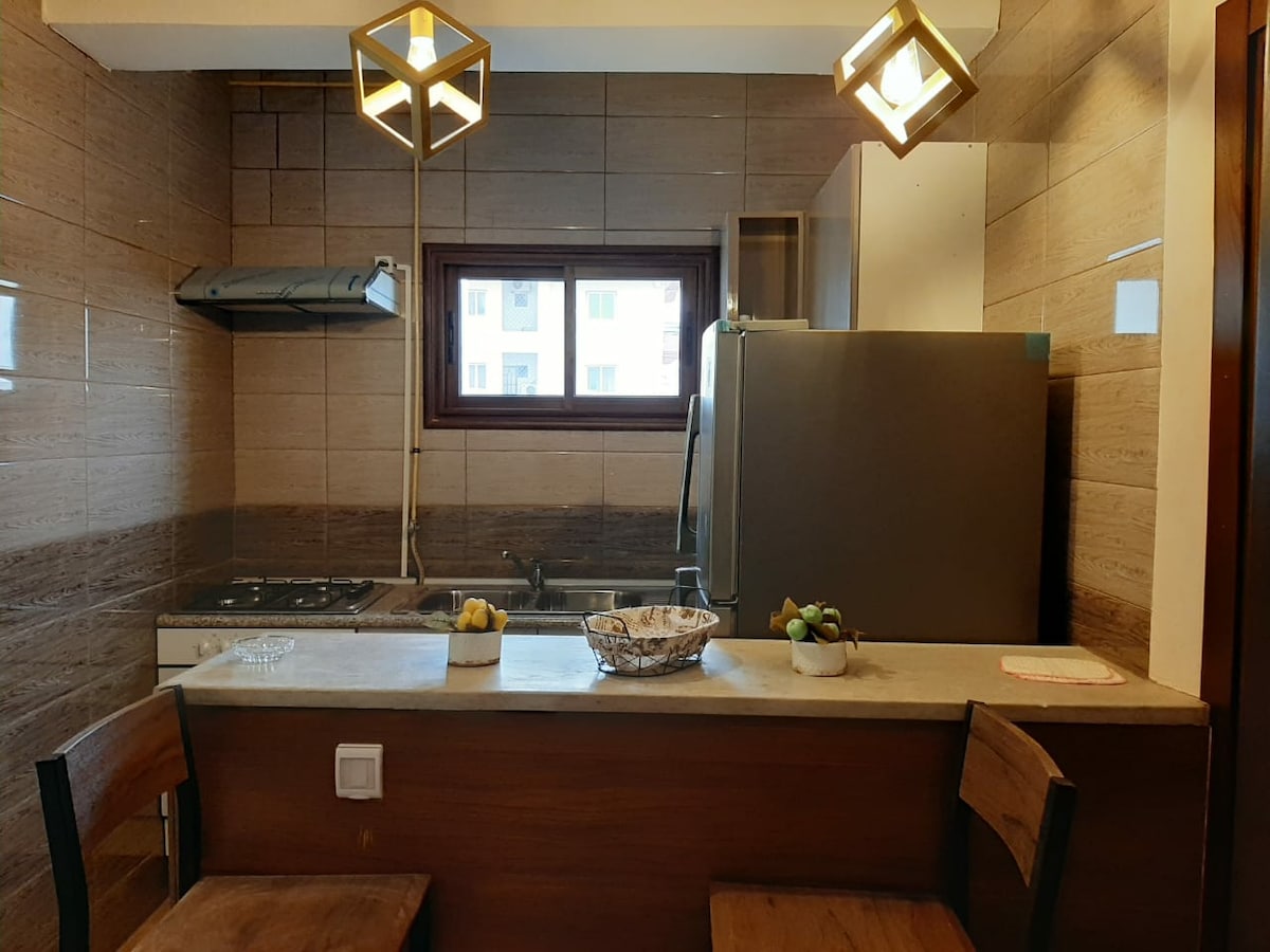 A modern kitchen space is featured, showcasing a large refrigerator and a stove against tiled walls. Two wooden bar stools are positioned at a counter, while light fixtures hang above. A window lets in natural light, illuminating a bowl of fruit on the counter.