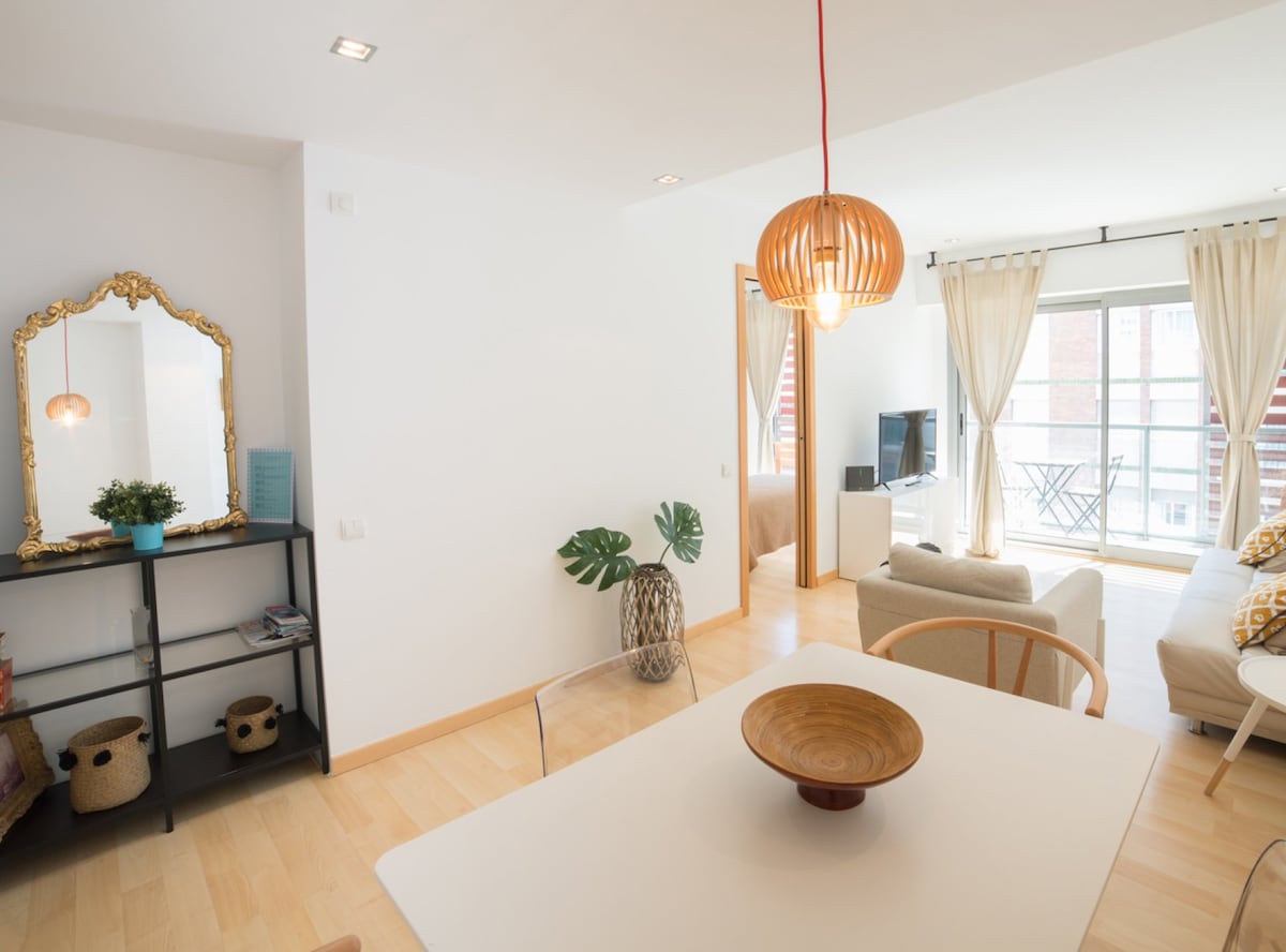 A bright living area is illuminated by natural light, featuring a modern wooden pendant lamp. A comfortable sofa and small side tables create a cozy setting, while a light wood dining table is complemented by a decorative bowl. A mirror and decorative plant add warmth to the space.