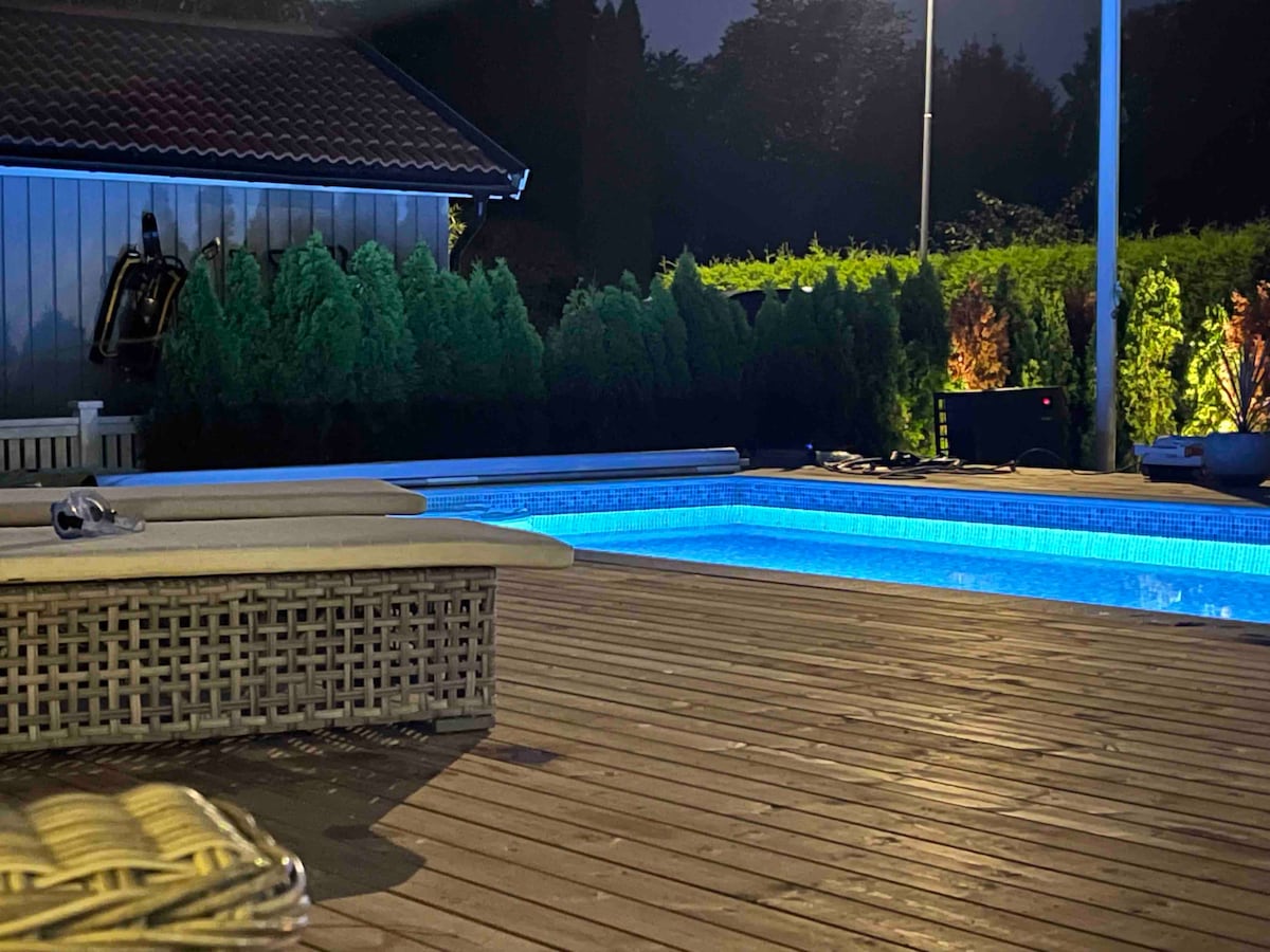 A heated swimming pool is illuminated with soft blue light, surrounded by wooden decking. Dense greenery lines the perimeter, adding privacy. Relaxing seating options are visible nearby, creating a welcoming outdoor space for guests.