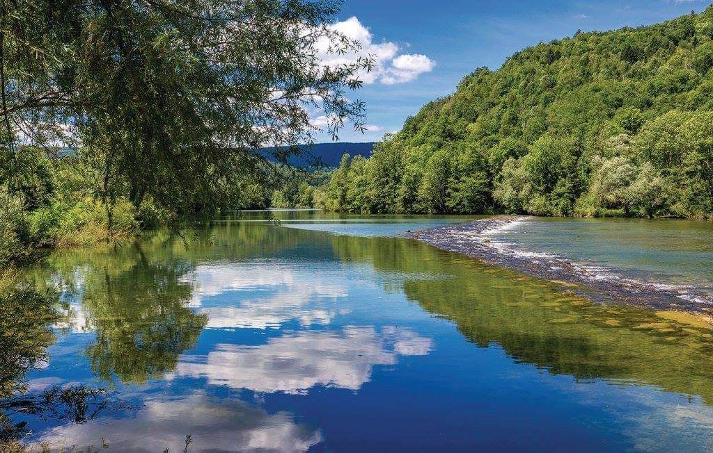 A calm river reflects lush green trees and a clear sky. Gentle ripples create a natural pattern on the water's surface, while clouds are mirrored vividly. The surrounding hillside is covered in greenery, contributing to a serene landscape that invites relaxation.