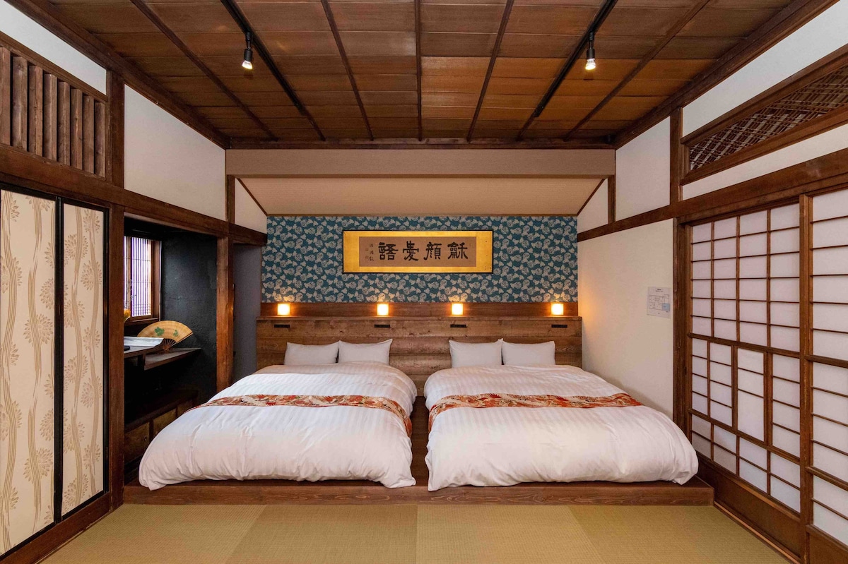 A traditional room features two spacious beds with neatly arranged bedding. The warm wooden walls are complemented by a decorative blue wallpaper. Sliding shoji doors provide flexibility to separate spaces, while ambient lighting creates a serene environment.