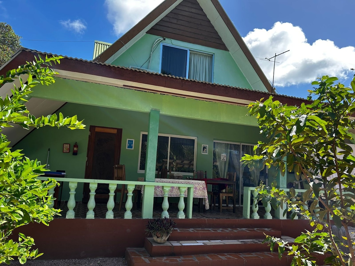 The exterior of the villa showcases a charming two-story structure with a sloping roof. Lush greenery surrounds the entrance, complemented by a welcoming porch with a wooden railing. Large windows allow natural light to illuminate the downstairs area, creating a cheerful ambiance.