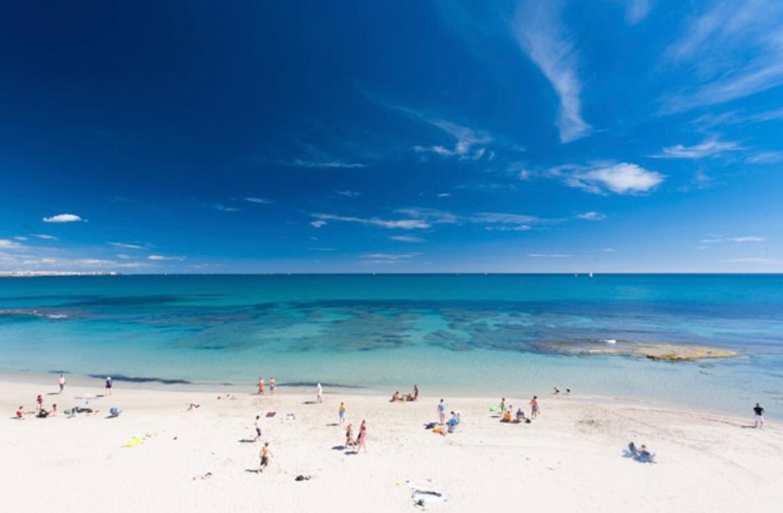 A sandy beach extends along the shoreline, where numerous individuals enjoy the sun and water. The vibrant turquoise sea contrasts with the clear blue sky, dotted with wispy clouds. Rocky formations are visible in the water, adding natural texture to the scene.