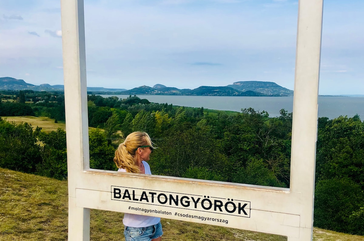 A young girl stands framed by a large white structure, overlooking the serene landscape of Lake Balaton. The horizon showcases gently rolling hills, while the expansive water reflects the sky. The surroundings feature lush greenery, illustrating a peaceful outdoor setting.
