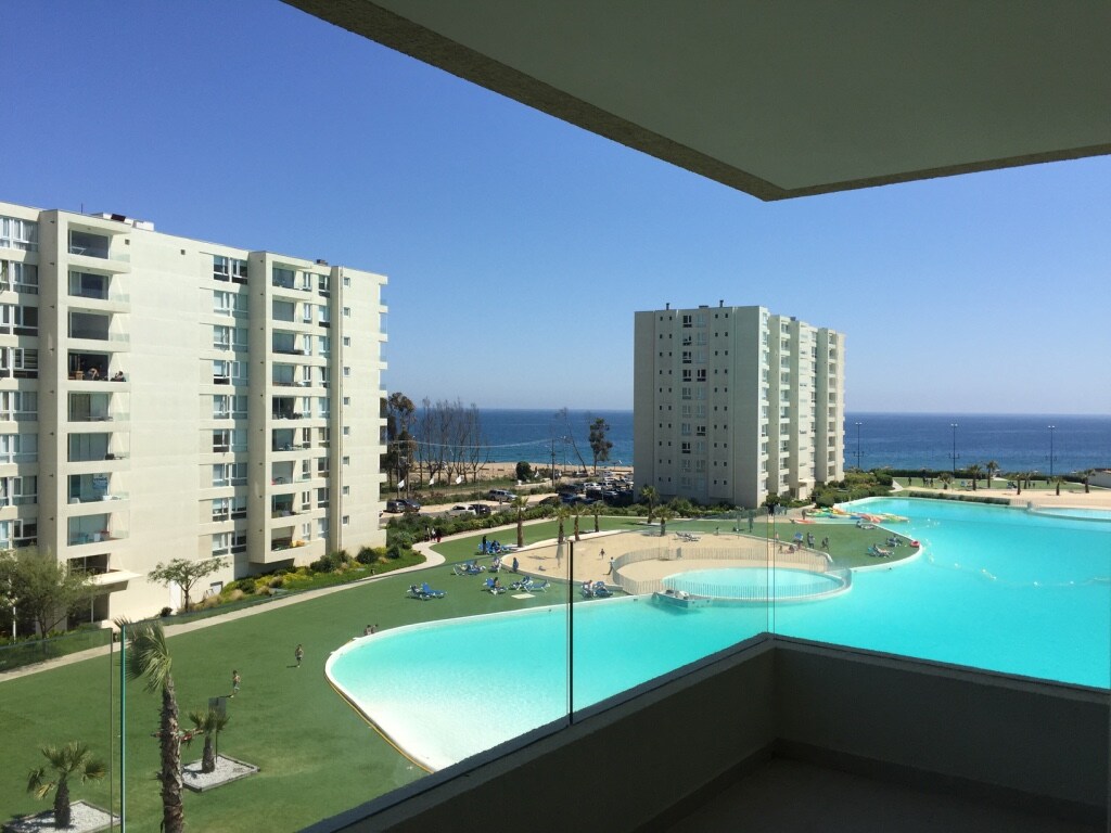 A scenic view is presented from the balcony, overlooking a large pool surrounded by green lawns and palm trees. Modern buildings are visible in the background, with the ocean gently glistening under clear blue skies.