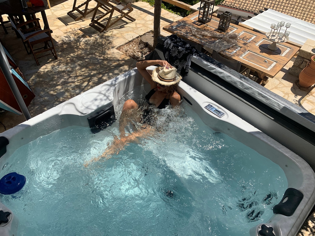 A hot tub is positioned outdoors, surrounded by natural stone tiles. Water is splashed as a person enjoys the experience, wearing a sun hat. Wooden chairs and a dining table are visible in the background, enhancing the outdoor space.