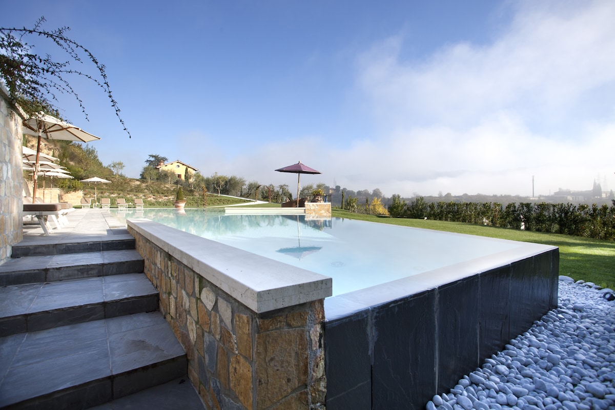 An inviting outdoor pool is showcased, featuring clear water that reflects the surrounding landscape. Comfortable loungers and shaded umbrellas are positioned nearby. The area is framed by greenery and a gentle slope that leads to distant hills, creating a peaceful ambiance.
