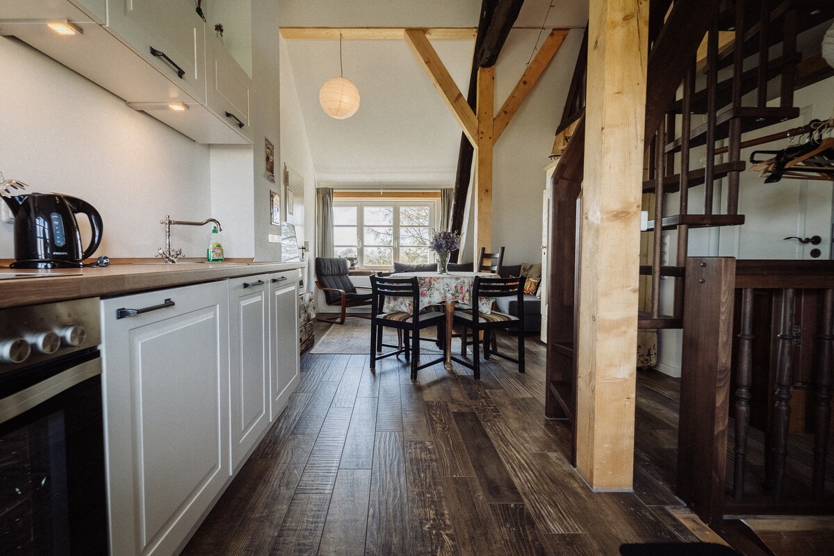 The open living area features a modern kitchen with white cabinetry and appliances. A wooden dining table with chairs sits near a window, allowing natural light to fill the space. Exposed wooden beams add character, and a staircase leads to the upper sleeping area.