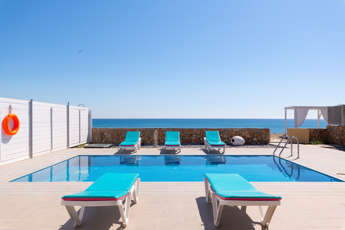 A private pool area is featured with turquoise lounge chairs arranged around a clear blue pool. Beyond the pool, the view extends over the calm sea under a clear sky, with a stone wall and sunshade providing a pleasant backdrop.