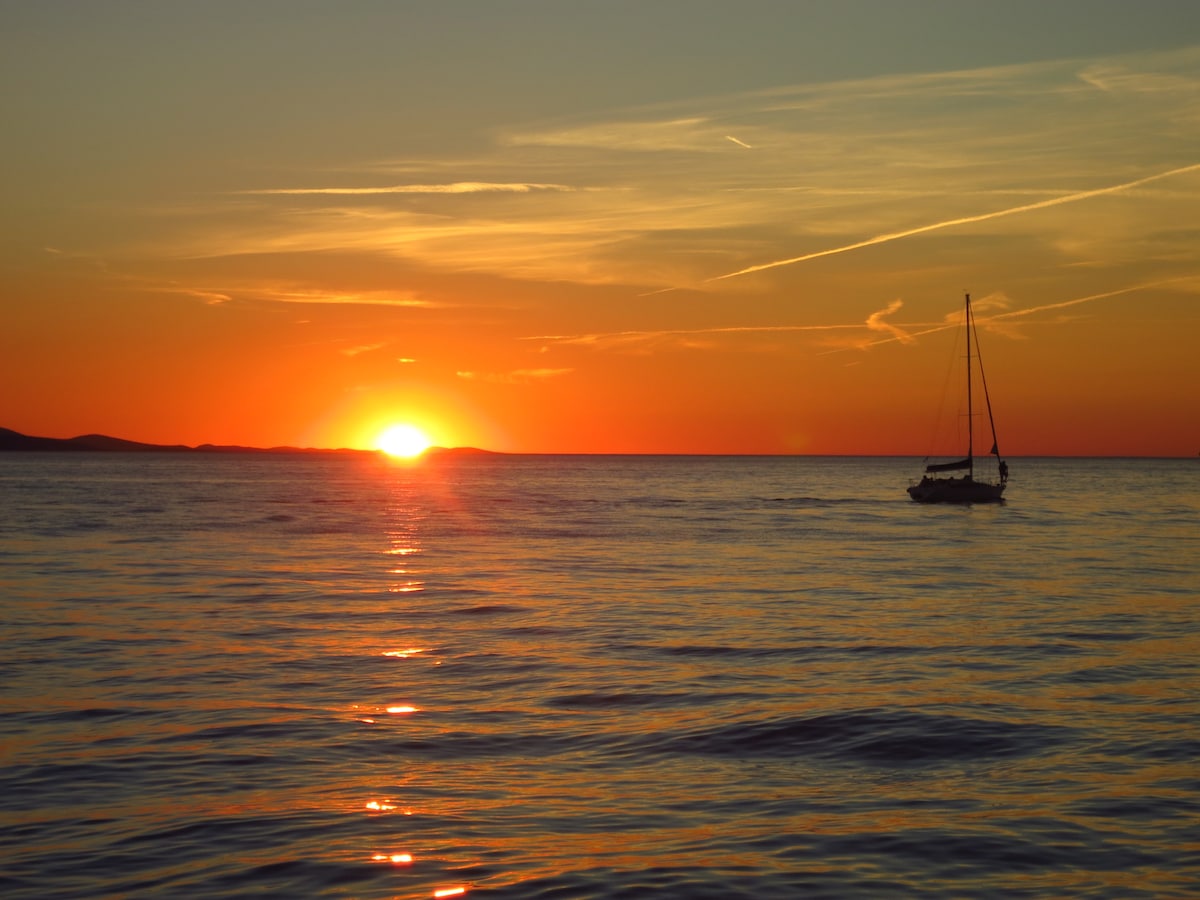 A colorful sunset is captured over the tranquil sea, with hues of orange, pink, and purple blending in the sky. A sailboat is visible, gently navigating the calm waters, while the sun gradually descends towards the horizon, creating a serene and picturesque seascape.