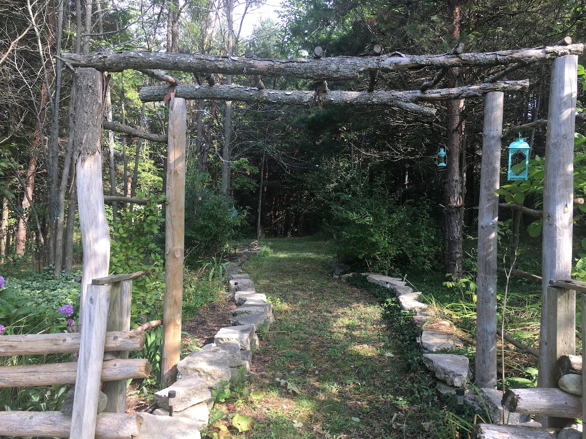 A rustic wooden archway frames a narrow path, flanked by stone borders leading into a forested area. Tall trees and greenery provide a natural canopy, creating a serene atmosphere. The ground is covered with grass, inviting peaceful exploration.