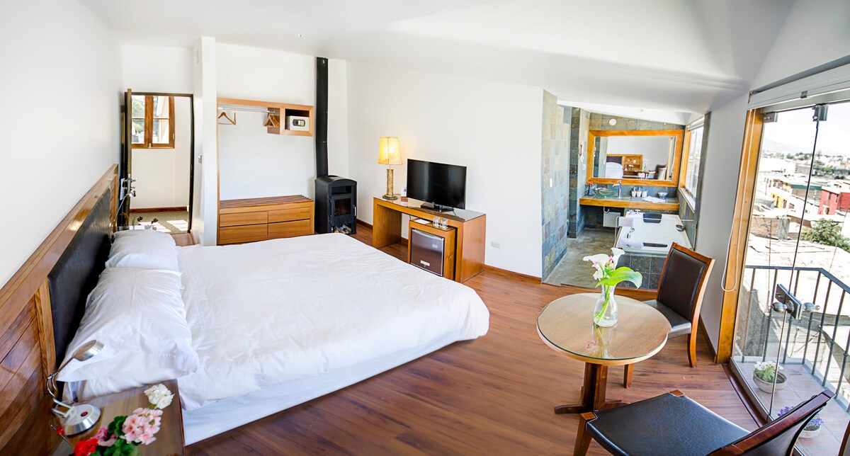 The room features a spacious bed dressed in white linens, complemented by a wooden headboard. A flat-screen television is mounted on the wall opposite. A small dining table is placed near a large window, which provides ample natural light. A glimpse of the modern bathroom can be seen.