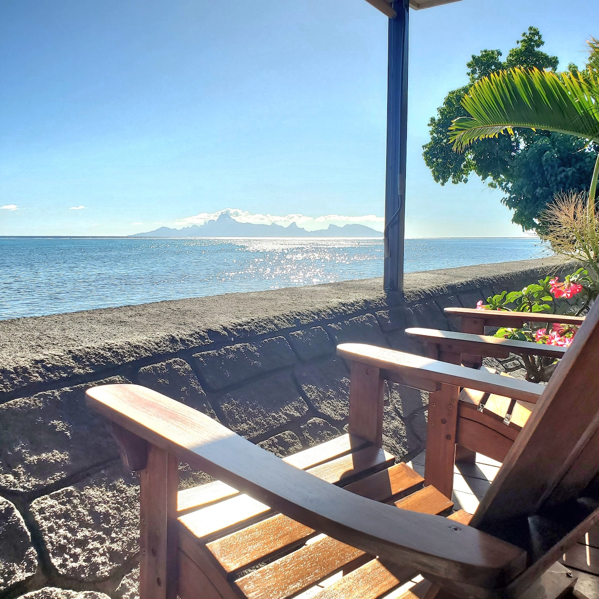 A wooden chair is positioned on a patio overlooking the serene water. The view includes distant mountains under a clear blue sky, while lush greenery and colorful flowers frame the scene. The gentle waves reflect sunlight, creating a calming atmosphere.