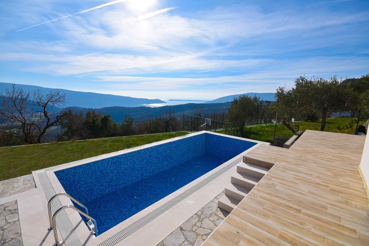 A private swimming pool is set against a backdrop of rolling hills and distant sea views. The pool features steps leading down into the water, and is surrounded by a wooden deck and manicured grass, providing a peaceful outdoor space.