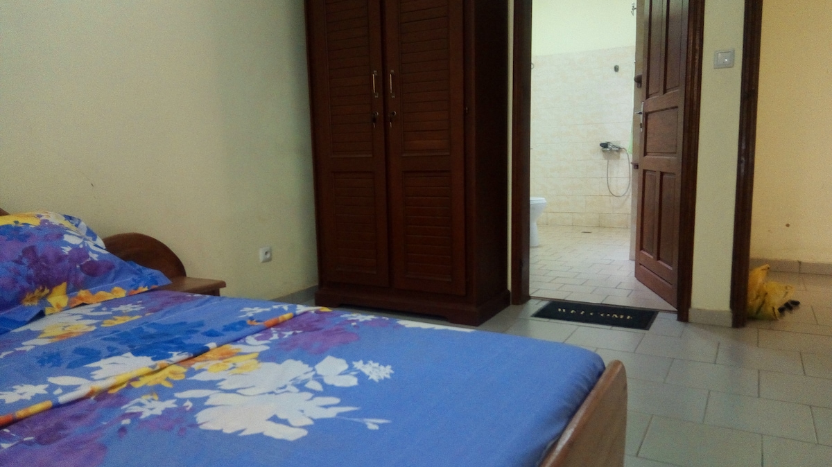 A spacious bedroom features a bed adorned with floral-patterned linens. A wooden wardrobe is positioned against the wall, and a doorway leads to a bathroom in the background with light-colored walls and tiled flooring.