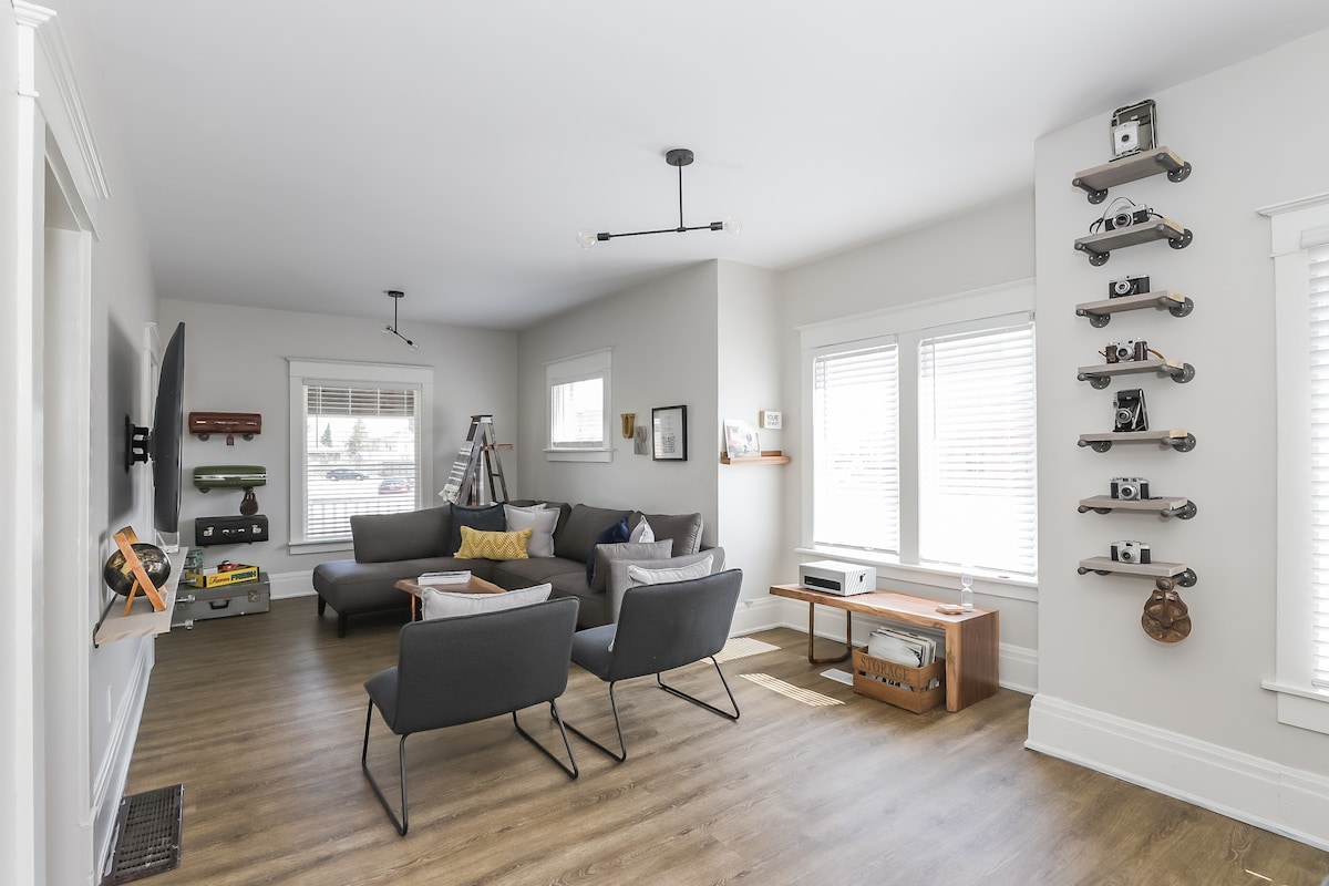 A modern living area features a spacious sectional sofa and two minimalist chairs arranged for conversation. Natural light enters through multiple windows with horizontal blinds. A wooden coffee table holds decor items, while a wall display showcases vintage cameras and art, enhancing the space's character.