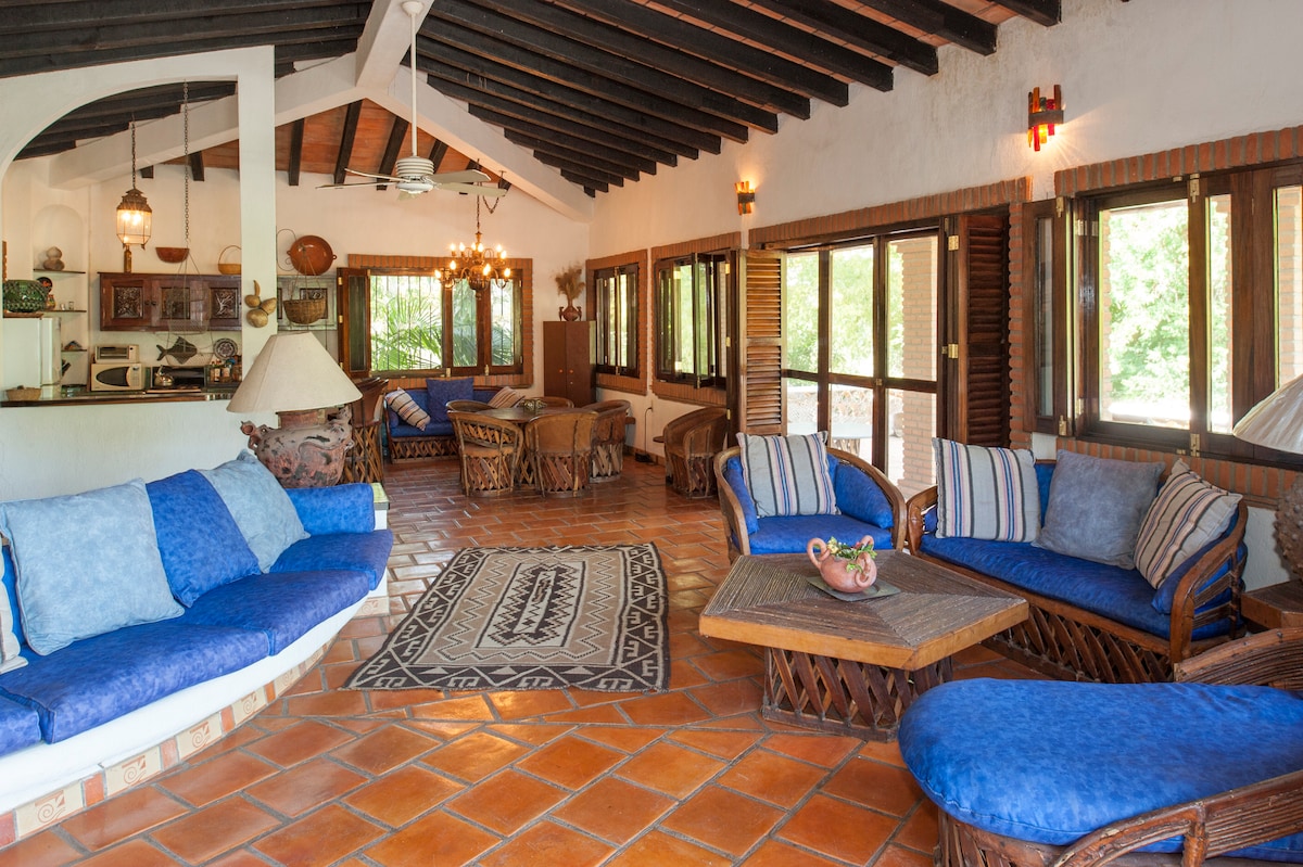 The living area features multiple seating arrangements, including blue upholstered sofas and wicker chairs. A large area rug sits on the terracotta-tiled floor, while natural light fills the space through wooden-framed windows, which open to views of the surrounding greenery.