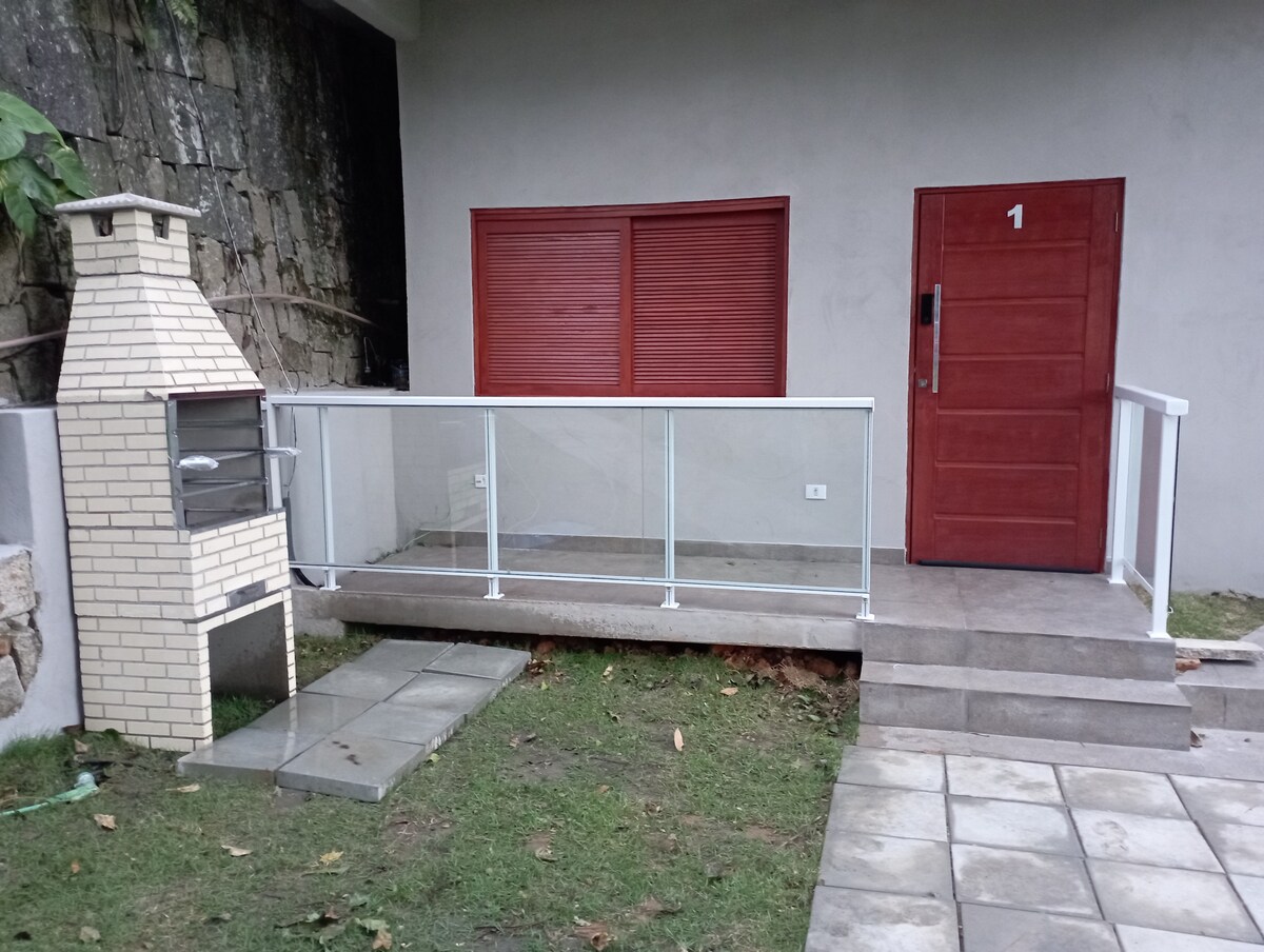The entrance to the chalet is framed by a clear glass railing, leading to a sturdy red door. Adjacent to the entrance, a brick barbecue grill is positioned, enhancing the outdoor space. A green lawn surrounds the area, providing a natural setting.