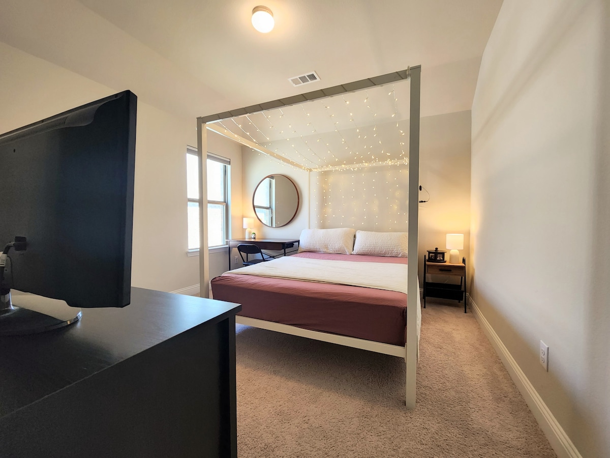 A well-lit bedroom features a spacious bed with a simple, elegant design, complemented by soft bedding. An entertainment unit is positioned near the foot of the bed, while a round mirror and bedside lamps add functionality. Soft lighting creates a cozy atmosphere.